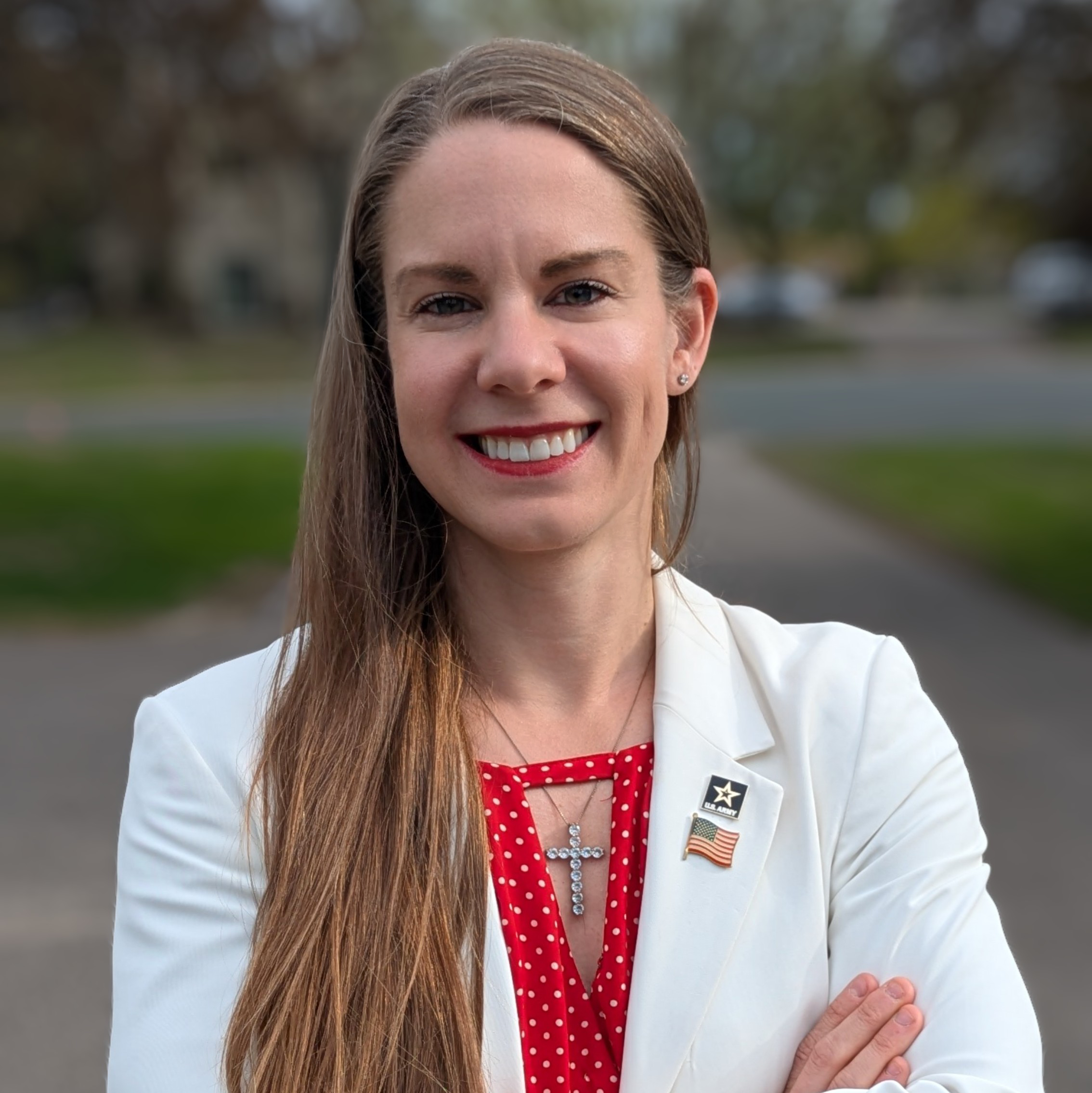 Portrait of Kristy Janigo, a white woman with long brown hair, wearing a white blazer over a red shirt, with a cross necklace, army pin, and American flag pin.
