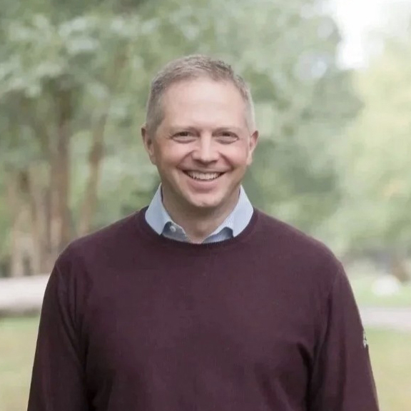 Portrait of Anders Folk, a middle-aged white man with short grey hair wearing a maroon sweater outside in front of green trees.