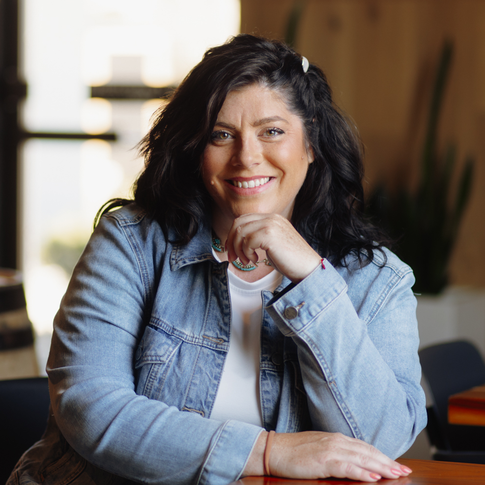 Portrait of Darci Smith, a white woman with black hair wearing a jean jacket over a white t-shirt in a coffee shop.