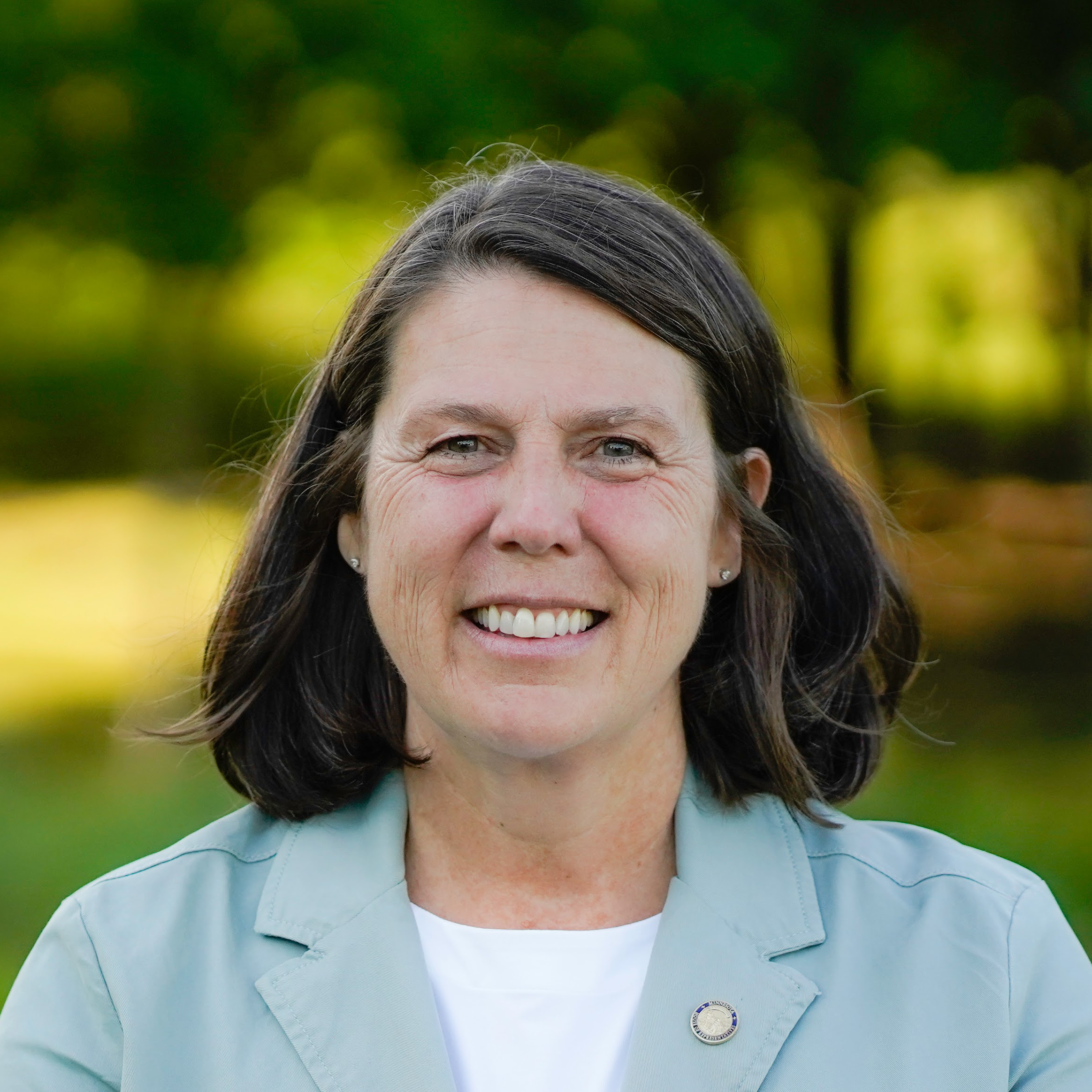 Portrait of Patty Acomb, a white woman with shoulder length brown hair wearing a light blue blazer, standing outside at golden hour.